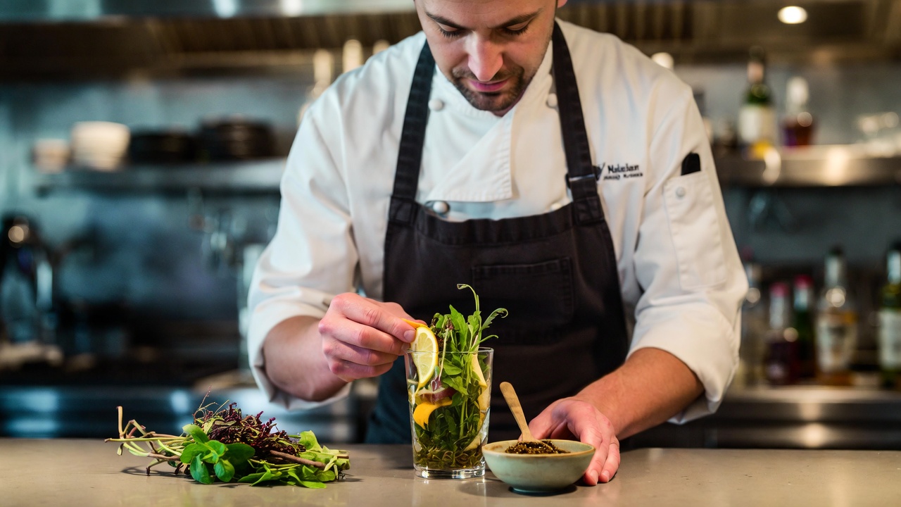 Chef in a Napa Valley restaurant preparing a zero proof beverage pairing using fresh herbs, citrus peel, and verjus for a sober travel tasting menu.
